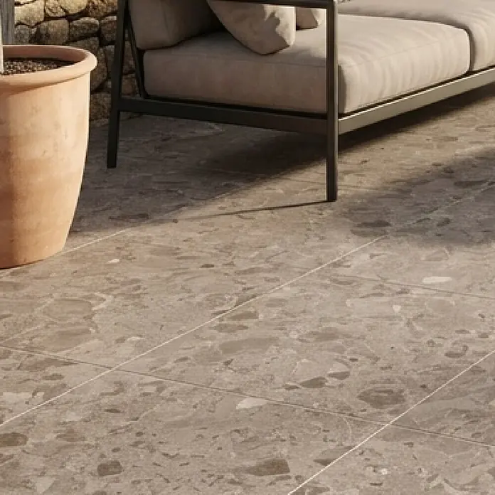Feinsteinzeugfliese Vogue - 60 x 60 cm, Brown, Matt - Große grau-braun gemusterte Outdoor-Bodenfliesen auf einer Terrasse, unter einem Sofa und neben einem Pflanzkübel verlegt.