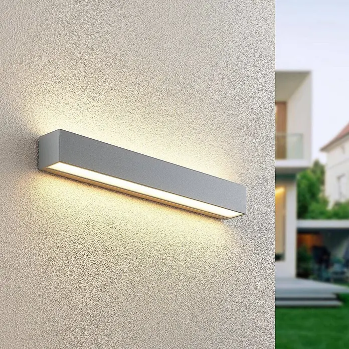 Lucande LED Wandleuchte aussen in silber, Aussenlampe Wand -  - Graue rechteckige Außenwandleuchte an strukturierter Wand, strahlt warmes Licht aus. Unscharfes modernes Haus und Garten im Hintergrund.