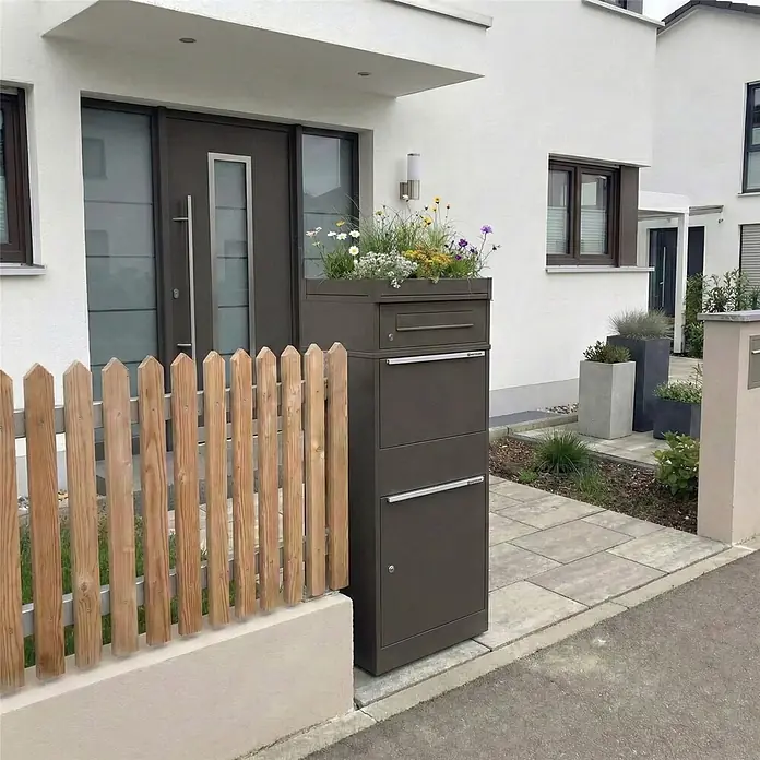 Metzler Paketbox BRFK PD BISPO - L x B x H: 37,4 x 50,3 x 119,9 cm, Eisenglimmer, Stahl, Paketfach, Mit Pflanzdach - Dunkelgrauer Paketkasten mit integriertem Pflanzgefäß, am Eingang eines modernen Hauses, neben einem Holzzaun.