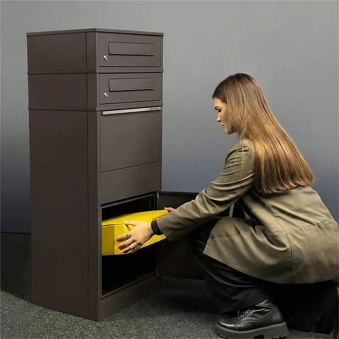 Metzler Paketbox 3 BRFK BISPO - L x B x H: 37,4 x 50,3 x 147,7 cm, Eisenglimmer, Stahl, Paketfach, 3 Briefkastenfächer - Dunkelbrauner Paketkasten mit mehreren Fächern. Eine Frau legt ein gelbes Paket in die untere Zustelltür.