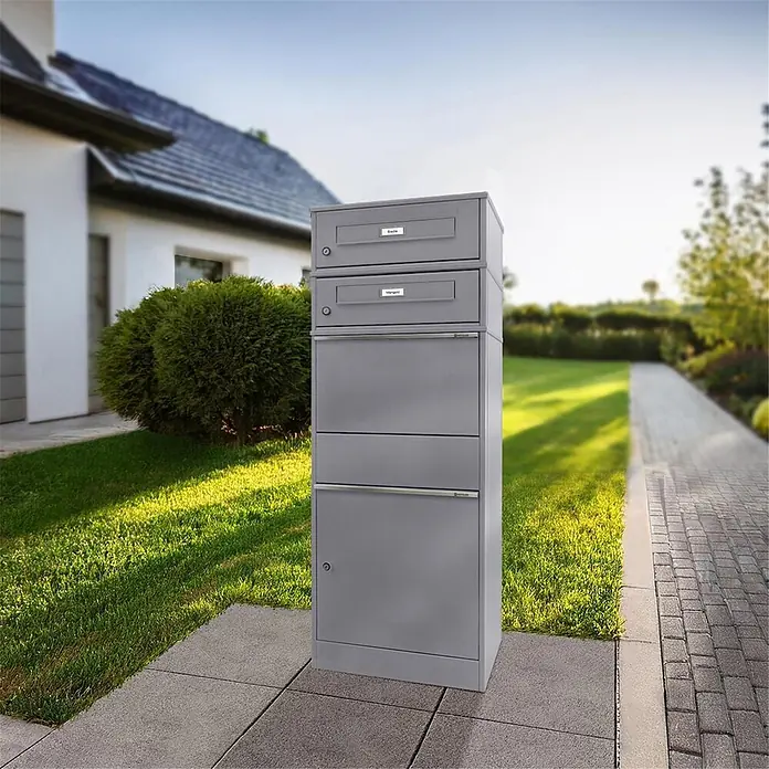 Metzler Paketbox 2 BRFK BISPO RE - L x B x H: 37,4 x 50,3 x 130,3 cm, Graualuminium, Stahl, Paketfach, Mit 2 Briefkastenfächern und Namensschildern - Eine hohe, graue, mehrteilige Brief- und Paketkastenanlage steht draußen auf einem gepflasterten Weg neben einem Haus.