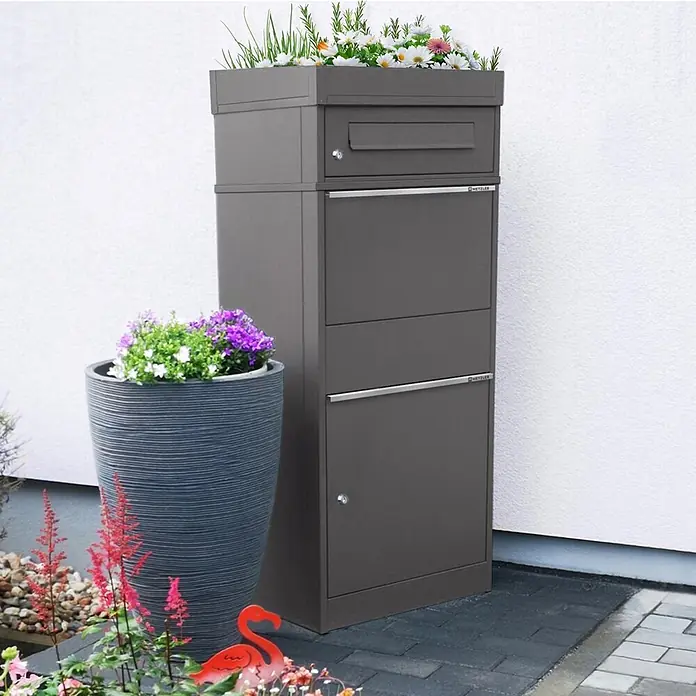 Metzler Paketbox BRFK PD BISPO - L x B x H: 37,4 x 50,3 x 119,9 cm, Eisenglimmer, Stahl, Paketfach, Mit Pflanzdach - Dunkelgrauer Paketbriefkasten mit Blumenkasten oben, draußen neben einem großen Blumentopf stehend.