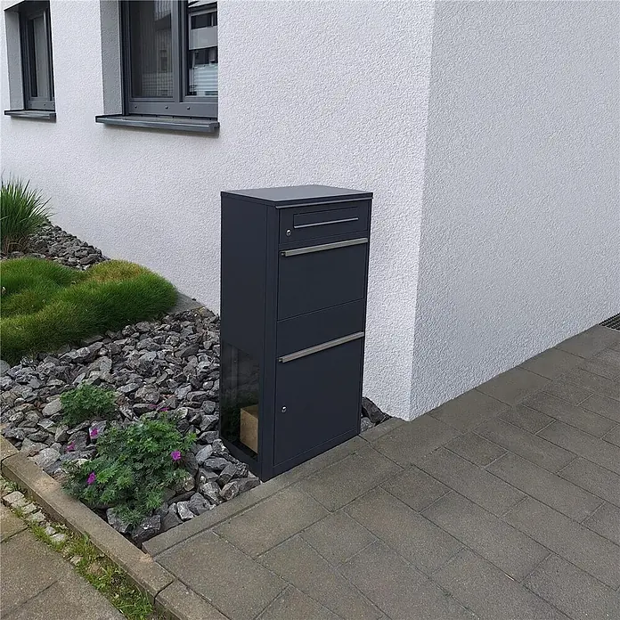 Metzler Paketbox BRFK BISPO SF - L x B x H: 37 x 50,2 x 112 cm, Anthrazit, Stahl, Paketfach, Mit Seitenfenster - Dunkelgraue Metall-Paketbox mit Briefschlitz und Paketfach, neben einem Haus platziert. Ein Paket ist innen sichtbar.