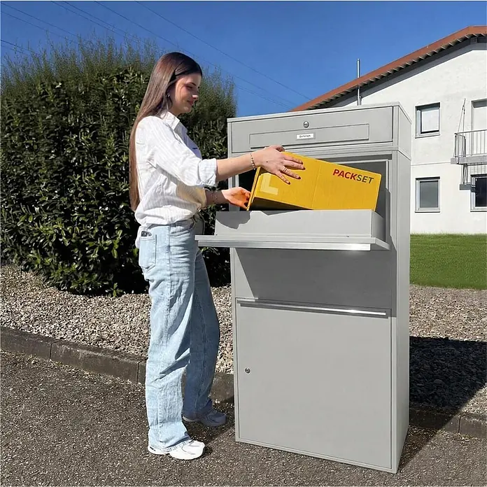 Metzler Paketbox XL BRFK MAX RE - L x B x H: 45,4 x 72 x 145 cm, Graualuminium, Stahl, Paketfach - Graue Paketbox in Gebrauch. Eine Frau legt ein gelbes Paket in die graue Metall-Paketbox.