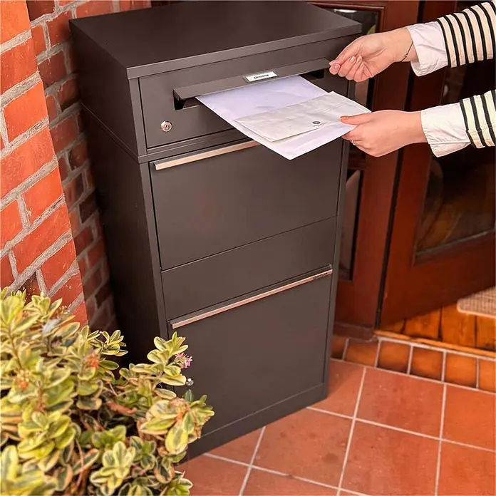 Metzler Paketbox BRFK BISPO RE - L x B x H: 37,4 x 50,3 x 112,9 cm, Eisenglimmer, Stahl, Paketfach - Dunkelbrauner Metall-Paketbriefkasten. Eine Hand legt ein Paket in den oberen Einwurfschlitz.