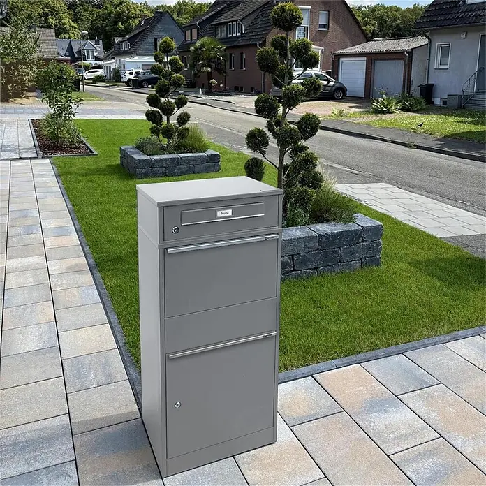 Metzler Paketbox BRFK BISPO RE - L x B x H: 37,4 x 50,3 x 112,9 cm, Graualuminium, Stahl, Paketfach - Hoher grauer Metallbriefkasten und Paketbox auf einem gepflasterten Weg in einer Wohngegend im Freien.