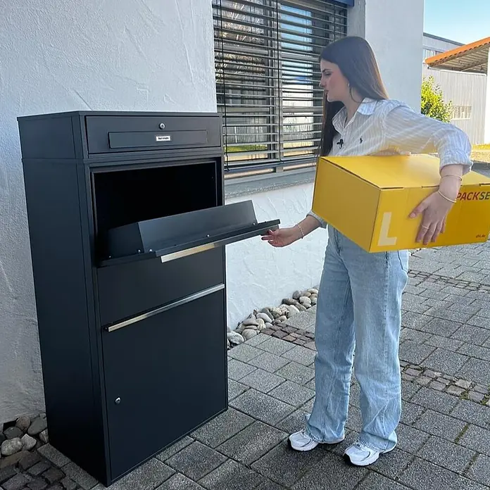 Metzler Paketbox XL BRFK MAX RE - L x B x H: 45,4 x 72 x 145 cm, Anthrazit, Stahl, Paketfach - Schwarze Paketbox aus Metall mit Briefkasten, gezeigt mit einer Person, die ein gelbes Paket einlegt.