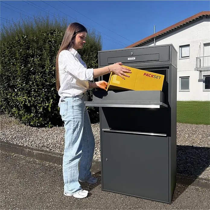 Metzler Paketbox XL BRFK MAX RE - L x B x H: 45,4 x 72 x 145 cm, Anthrazit, Stahl, Paketfach - Dunkelgrauer Paketkasten zur sicheren Paketzustellung, gezeigt mit einer Person, die ein gelbes Paket einlegt.