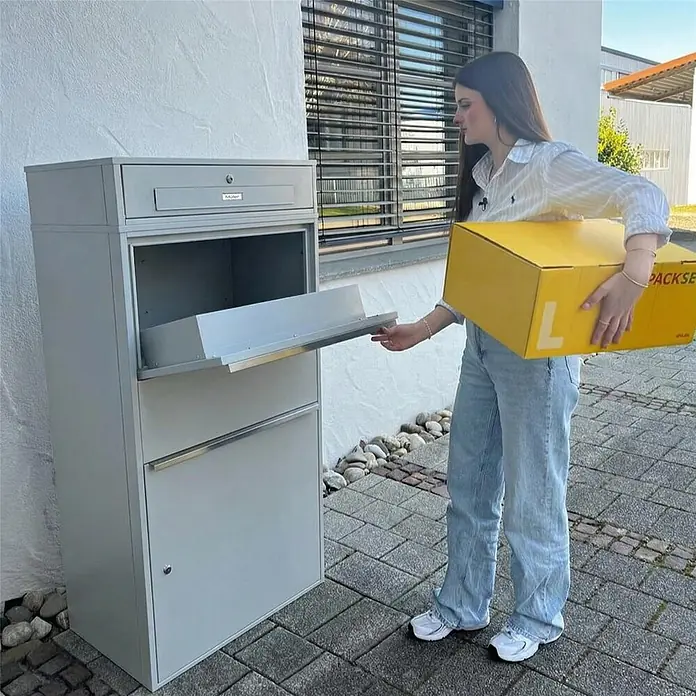 Metzler Paketbox XL BRFK MAX RE - L x B x H: 45,4 x 72 x 145 cm, Graualuminium, Stahl, Paketfach - Graue Paketbox, in die eine Frau ein gelbes Paket legt, was ihre Nutzung zur sicheren Paketzustellung zeigt.