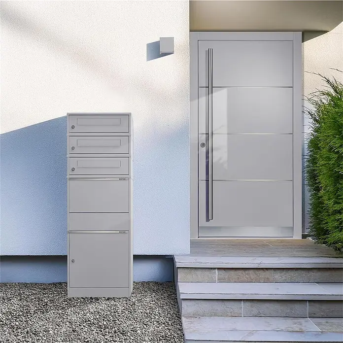Metzler Paketbox 3 BRFK BISPO - L x B x H: 37,4 x 50,3 x 147,7 cm, Graualuminium, Stahl, Paketfach, 3 Briefkastenfächer - Hohes, graues Mehrfachbriefkastensystem auf Kies neben einer modernen grauen Haustür.