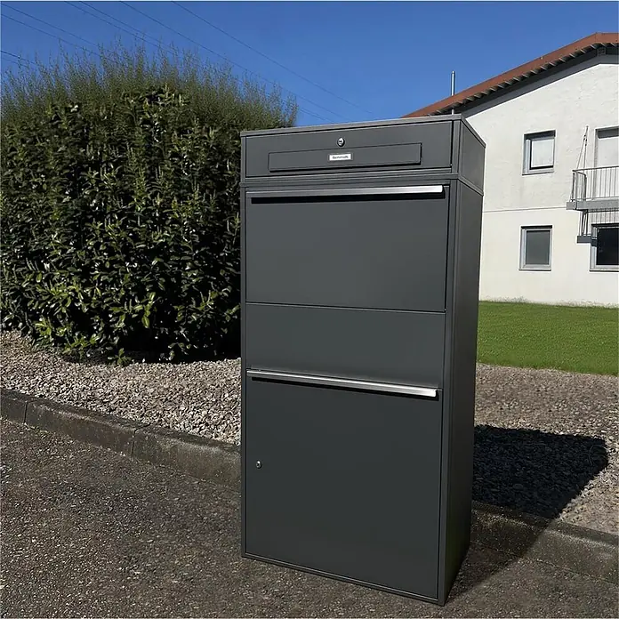 Metzler Paketbox XL BRFK MAX RE - L x B x H: 45,4 x 72 x 145 cm, Anthrazit, Stahl, Paketfach - Dunkelgrauer freistehender Paketbriefkasten mit mehreren Fächern, draußen neben einem Busch und einem gepflasterten Weg.