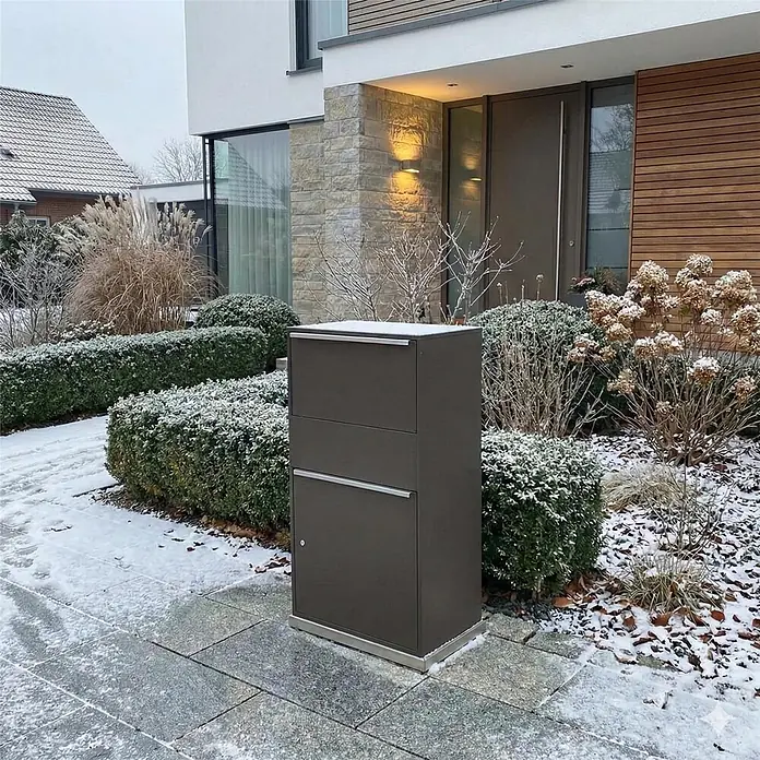 Metzler Paketbox XL BISPO MAX - L x B x H: 45,4 x 72 x 128,5 cm, Eisenglimmer, Stahl, Paketfach - Dunkelgrauer Metall-Paketbriefkasten auf gepflastertem Weg vor einem modernen Haus, umgeben von schneebedeckten Winterpflanzen.