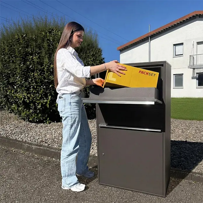 Metzler Paketbox XL BISPO MAX - L x B x H: 45,4 x 72 x 128,5 cm, Eisenglimmer, Stahl, Paketfach - Eine Person legt ein gelbes 'PACKSET'-Paket in eine dunkelgraue, moderne Paketbox.