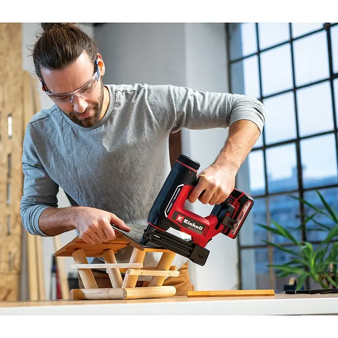 Einhell PXC 18V Akku-Nagler - 18 V, Ohne Akku, Länge Nagel: 15 mm - 32 mm - Mann verwendet roten und schwarzen Akku-Nagler, um Material an einem Holz-Vogelhaus zu befestigen.