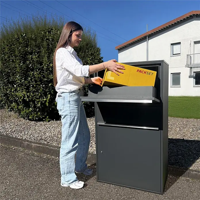 Metzler Paketbox XL BISPO MAX - L x B x H: 45,4 x 72 x 128,5 cm, Anthrazit, Stahl, Paketfach - Dunkelgraue Paketbox für den Außenbereich, in die eine Frau ein gelbes Paket einlegt.