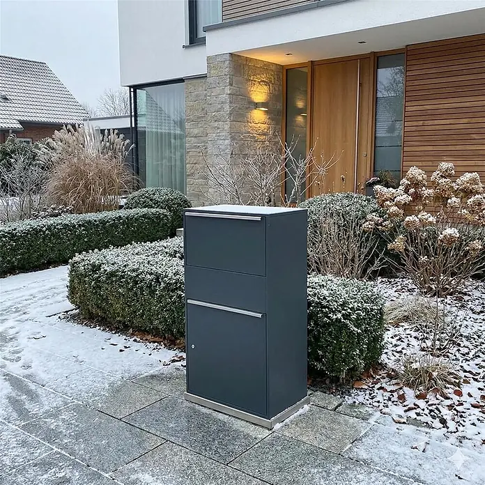 Metzler Paketbox XL BISPO MAX - L x B x H: 45,4 x 72 x 128,5 cm, Anthrazit, Stahl, Paketfach - Dunkelgrauer moderner Paketbriefkasten mit Edelstahlsockel in verschneitem Vorgarten vor einem Haus.