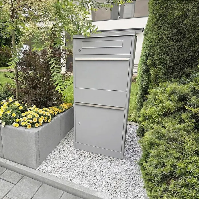 Metzler Paketbox BRFK BISPO - L x B x H: 37,4 x 50,3 x 112,9 cm, Graualuminium, Stahl, Paketfach - Hoher, grauer Brief- und Paketkasten aus Metall, steht im Freien auf weißen Kieselsteinen, mit mehreren Fächern für Post und Pakete.