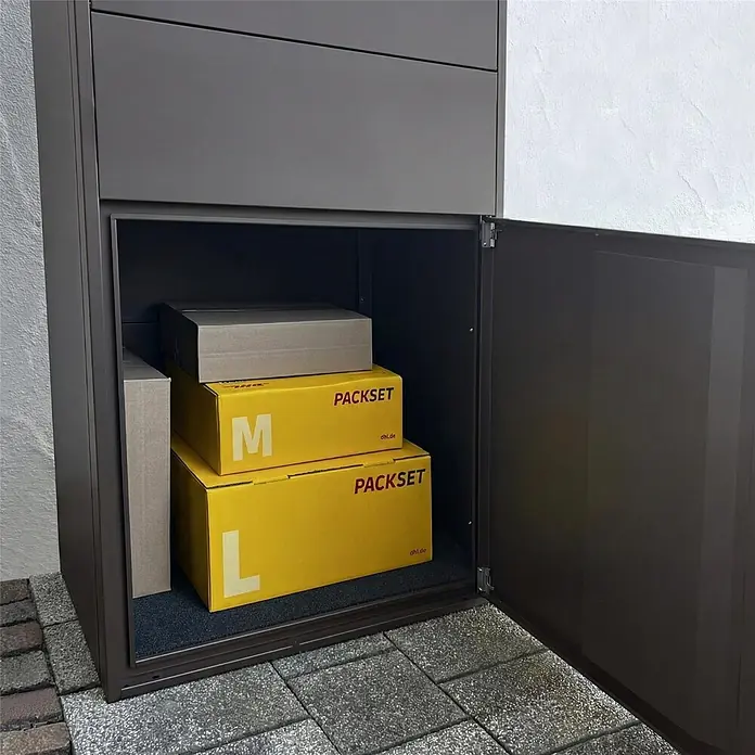 Metzler Paketbox XL BRFK BISPO MAX - L x B x H: 45,4 x 72 x 145 cm, Eisenglimmer, Stahl, Paketfach - Dunkelgraue Metall-Paketbox, Tür offen, mit gelben und braunen Paketen. Draußen auf Pflastersteinen installiert.