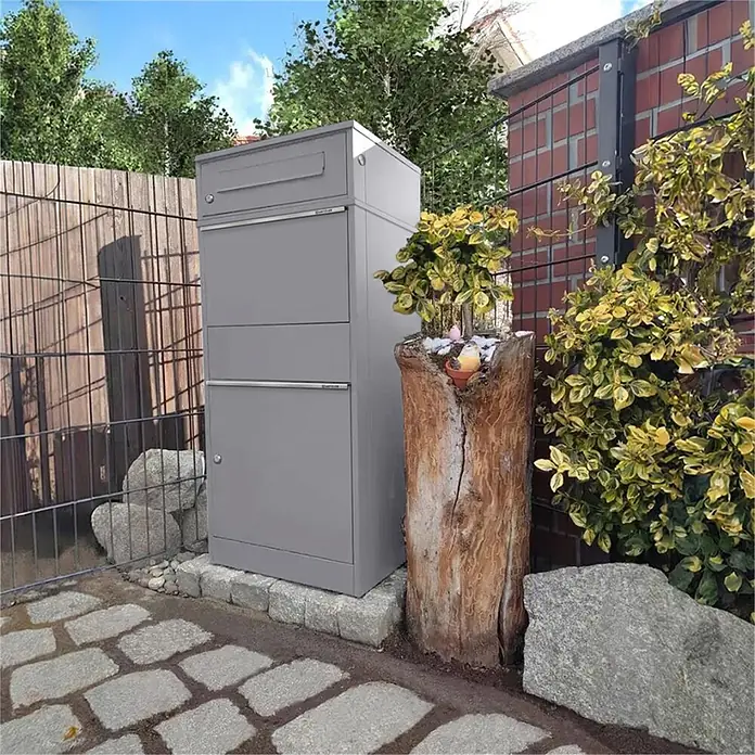 Metzler Paketbox BRFK BISPO - L x B x H: 37,4 x 50,3 x 112,9 cm, Graualuminium, Stahl, Paketfach - Ein hoher, grauer Paketbriefkasten aus Metall steht draußen auf Pflastersteinen, neben einem Holzzaun und einer Ziegelmauer.