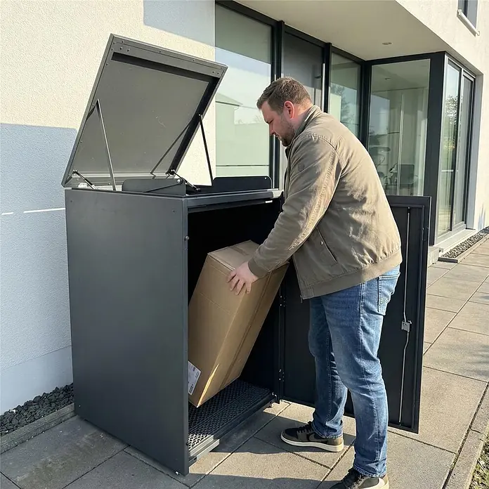 Metzler Paketbox XXL BEN - L x B x H: 81,2 x 68,4 x 123,3 cm, Anthrazit, Stahl - Ein Mann legt ein großes Paket in eine dunkelgraue, freistehende Paketbox vor einem modernen Gebäude.