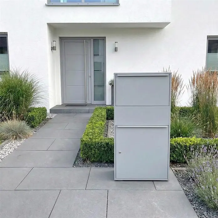 Metzler Paketbox XL BISPO MAX - L x B x H: 45,4 x 72 x 128,5 cm, Graualuminium, Stahl, Paketfach - Moderner, großer grauer Paketbriefkasten mit mehreren Fächern, steht auf einem gepflasterten Weg vor einem Haus.
