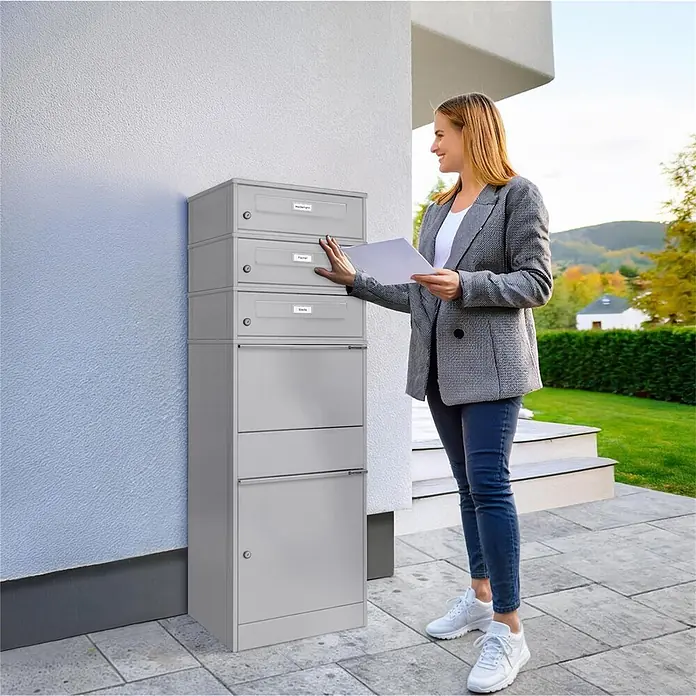 Metzler Paketbox 3 BRFK BISPO RE - L x B x H: 37,4 x 50,3 x 147,7 cm, Graualuminium, Stahl, Namensschild, 3 Briefkastenfächer und Namensschildern - Ein hohes, graues Mehrfachanlagen-Briefkastensystem aus Metall, das von einer Frau benutzt wird, die Post in ein oberes Fach neben einem Haus steckt.