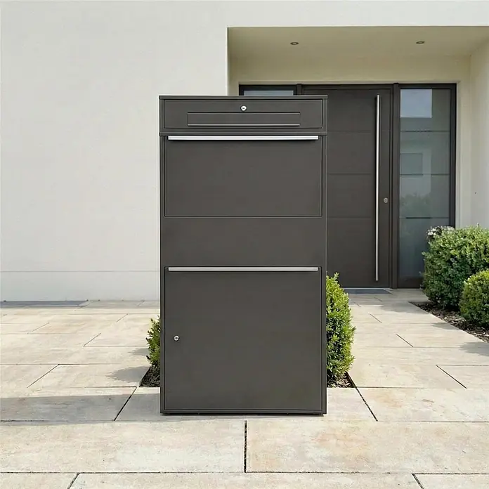 Metzler Paketbox XL BRFK BISPO MAX - L x B x H: 45,4 x 72 x 145 cm, Eisenglimmer, Stahl, Paketfach - Dunkelgrauer moderner Brief- und Paketkasten mit silbernen Akzenten, auf gepflastertem Weg vor einem Haus. Mehrere Fächer für Post und Pakete.
