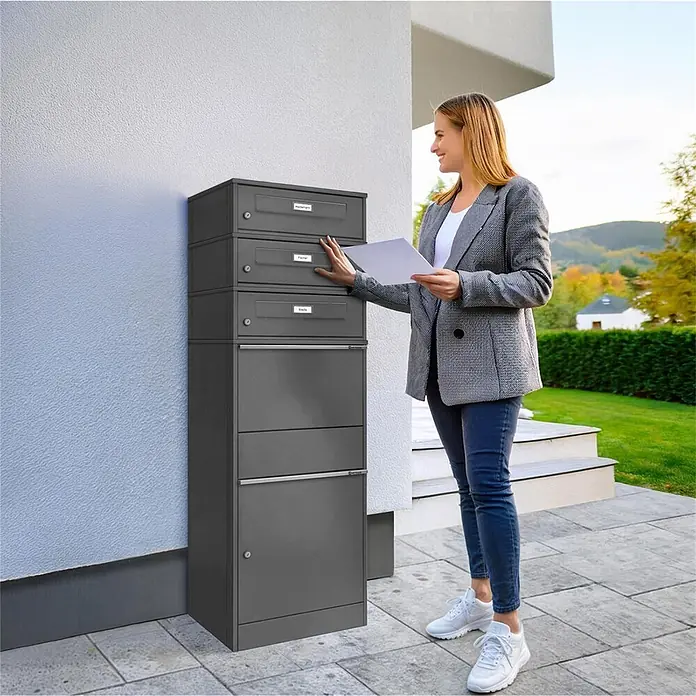 Metzler Paketbox 3 BRFK BISPO RE - L x B x H: 37,4 x 50,3 x 147,7 cm, Eisenglimmer, Stahl, Namensschild, 3 Briefkastenfächer und Namensschildern - Dunkelgraue Paketbox mit mehreren Fächern, neben einem Haus, eine Frau entnimmt Post.