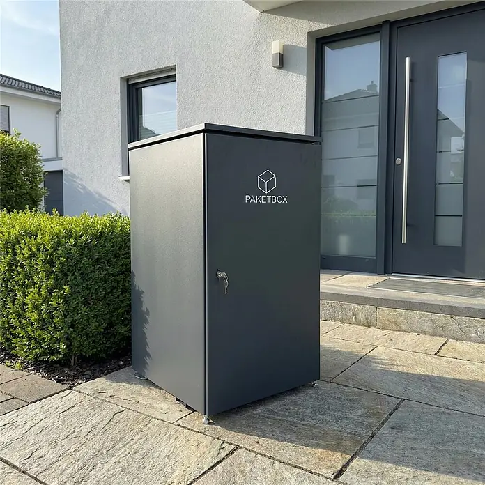 Metzler Paketbox XXL BEN - L x B x H: 81,2 x 68,4 x 123,3 cm, Anthrazit, Stahl - Dunkelgraue Paketbox mit Schlüssel und Würfellogo, auf einer Steinterrasse vor einem modernen Haus platziert.