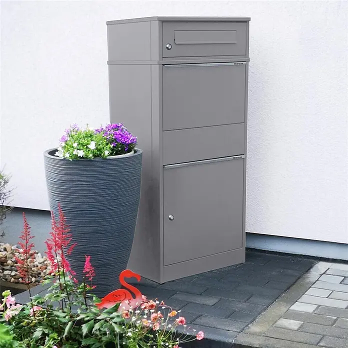 Metzler Paketbox BRFK BISPO - L x B x H: 37,4 x 50,3 x 112,9 cm, Graualuminium, Stahl, Paketfach - Hoher grauer Metall-Paketbriefkasten steht neben einem Hauseingang, mit Pflanzgefäß und Pflanzen daneben.