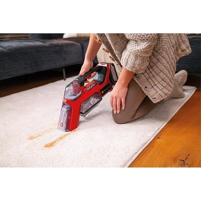 Einhell PXC 18V Akku-Waschsauger TE-SCL 18/200 Li-Solo - 18 V, Ohne Akku, Behältervolumen: 0,2 l - Eine Person benutzt einen rot-schwarzen Hand-Fleckreiniger, um einen Fleck von einem weißen Teppich zu entfernen.