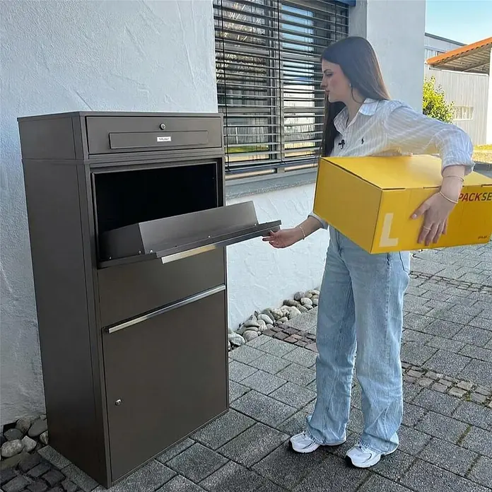Metzler Paketbox XL BRFK MAX RE - L x B x H: 45,4 x 72 x 145 cm, Eisenglimmer, Stahl, Paketfach - Dunkelbrauner Paketbriefkasten mit "Müller"-Branding. Eine Frau legt ein gelbes Paket in das geöffnete Paketfach.