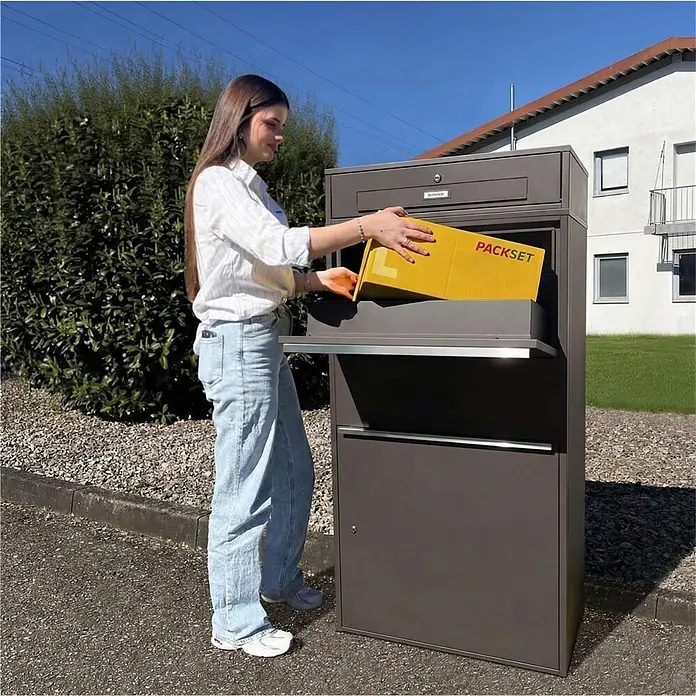 Metzler Paketbox XL BRFK MAX RE - L x B x H: 45,4 x 72 x 145 cm, Eisenglimmer, Stahl, Paketfach - Dunkelgrauer Paketkasten, Frau legt gelbes Paket in den Einwurfschlitz.