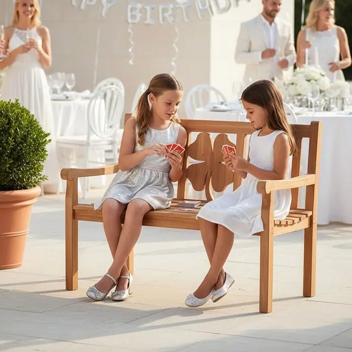 Gartenbank Kleeblatt Teak 120cm -  - Hölzerne Gartenbank mit dekorativer Kleeblatt-Rückenlehne, auf der zwei Kinder bequem Karten spielen bei einer Outdoor-Veranstaltung.