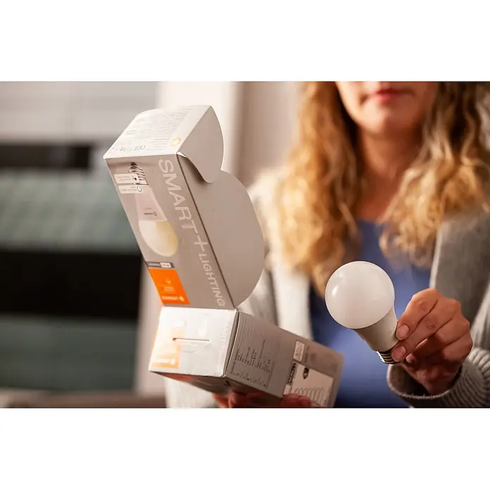 Osram LED-Leuchtmittel SMART+ MATTER Classic A100 14W 827...865 TW E27 - E27, Dimmbar, Mehrfarbig, 1.521 lm, 14 W - Eine Person hält eine weiße Smart-Glühbirne und deren offene Verpackung mit der Aufschrift 'SMART+ LIGHTING'.