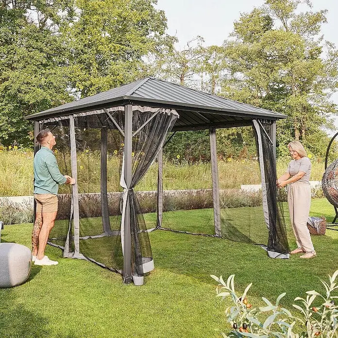 Home Deluxe Standard-Pavillon AZUR - Farbe Gestell: Anthrazit, Metall - Ein dunkelgrauer Pavillon mit schwarzem Moskitonetz im Garten. Zwei Personen richten das Netz aus.