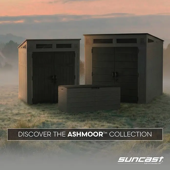 Suncast Auflagenbox - 150 x 61 x 64 cm, Kunststoff, Anthrazit, Geteilter Deckel - Zwei dunkelgraue Suncast Ashmoor Lagerschuppen und eine passende Kiste auf Gras in nebliger Naturkulisse.
