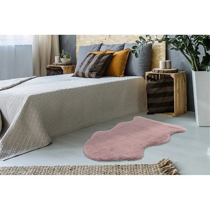 Kayoom Kunstfell Cosy Sheepskin 525 - Puderrosa, 90 x 60 cm, 100 % Polyester - Unregelmäßig geformter, flauschiger rosa Teppich auf dem Boden neben einem Bett.