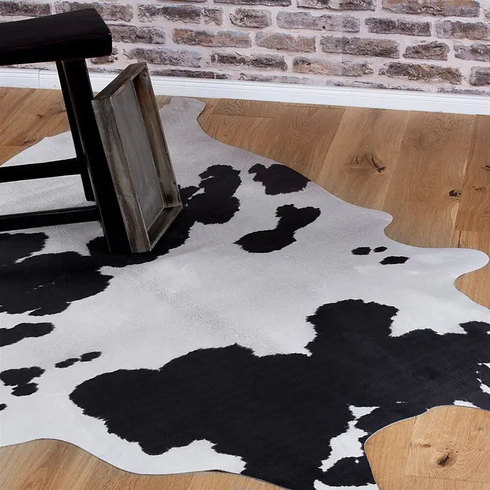 Kayoom Kunstfell Sanari 200 - Schwarz/Weiß, 190 x 155 cm, 100 % Polyester - Schwarz-weißer Kuhfellteppich mit Naturmuster auf hellem Holzboden.