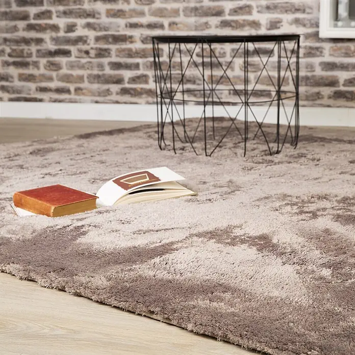 Kayoom Hochflorteppich Dolphin 500 - Taupe, 110 x 60 cm, 100 % Polyester - Ein zweifarbiger brauner Zottelteppich mit Marmormuster auf einem Holzfußboden, mit Büchern und einem Drahttisch.