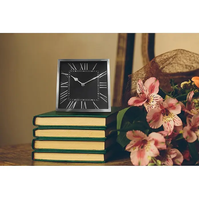 Tischuhr Moments 2025 - Silber, 21 x 21 cm - Eine quadratische schwarz-silberne Uhr mit römischen Ziffern steht auf grünen Büchern neben rosa Blumen.