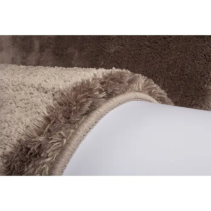 Kayoom Hochflorteppich Dolphin 500 - Taupe, 110 x 60 cm, 100 % Polyester - Nahaufnahme eines zweifarbigen, beigefarbenen und braunen, flauschigen Hochflorteppichs, teilweise gerollt, zeigt die weiche Textur und den genähten Rand.