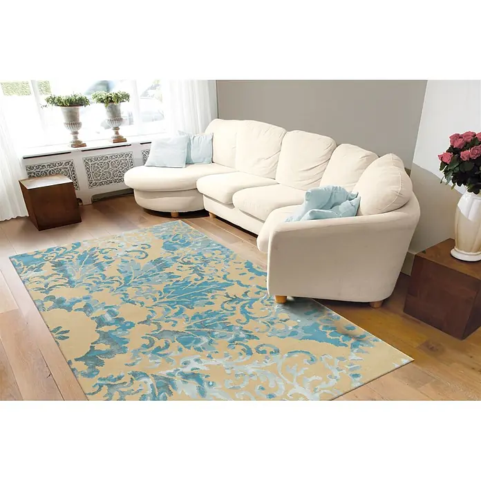 Kayoom Kurzflorteppich Satin 8061 - Creme/Türkis, 180 x 120 cm, 70 % Wolle, 30 % Viskose - Ein beigefarbener Teppich mit blauem, ornamentalem Muster auf Holzboden in einem Wohnzimmer, teilweise unter einem hellen Sofa.