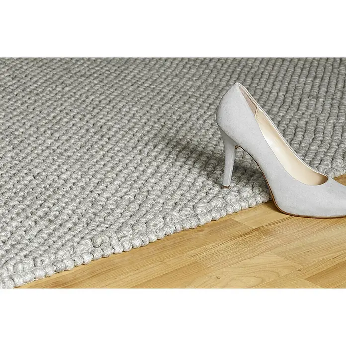 Kayoom Wollteppich Attic 590 - Silber, 170 x 120 cm, 50 % Wolle, 50 % Viskose - Ein hellgrauer, grob gestrickter Teppich mit einem Absatzschuh auf Holzboden.