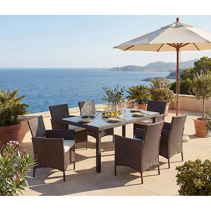 Mendler Poly-Rattan-Garnitur Halden - braun, Kissen Creme -  - Braunes Rattan-Garten-Essset mit Glastisch und hellen Kissen auf einer Terrasse mit Meerblick.