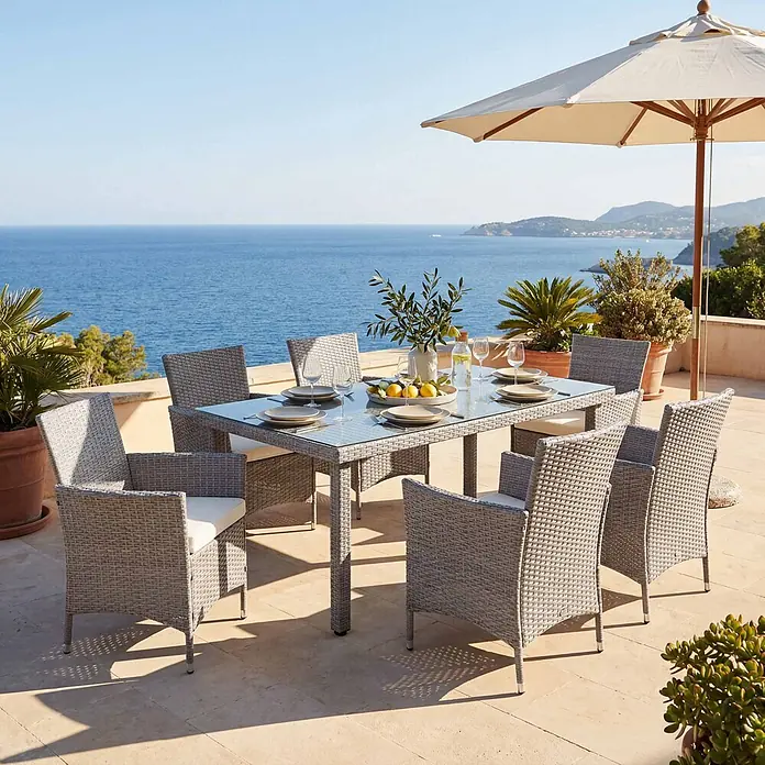 Mendler Gartenmöbel-Set - 7 -tlg., Polyrattan, Grau, Kissen creme, Grau, Kissen creme - Graues Rattanspeiseset mit Glastisch und Stühlen mit weißen Kissen auf einer sonnigen Terrasse mit Meerblick.
