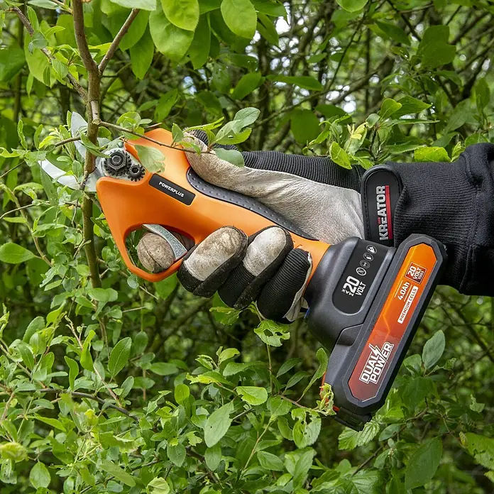 PowerPlus Tijeras para podar de batería + Motosierra - 20 V, 1 batería, 2 Ah, Grosor de rama máx.: 30 mm - Una mano enguantada usa una tijera de podar inalámbrica naranja y negra para cortar una rama verde.