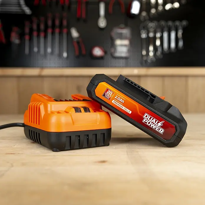 PowerPlus Tijeras para podar de batería + Motosierra - 20 V, 1 batería, 2 Ah, Grosor de rama máx.: 30 mm - Un cargador de batería naranja y negro para herramientas eléctricas con una batería de 20V, 2.0Ah a juego, sobre un banco de taller.