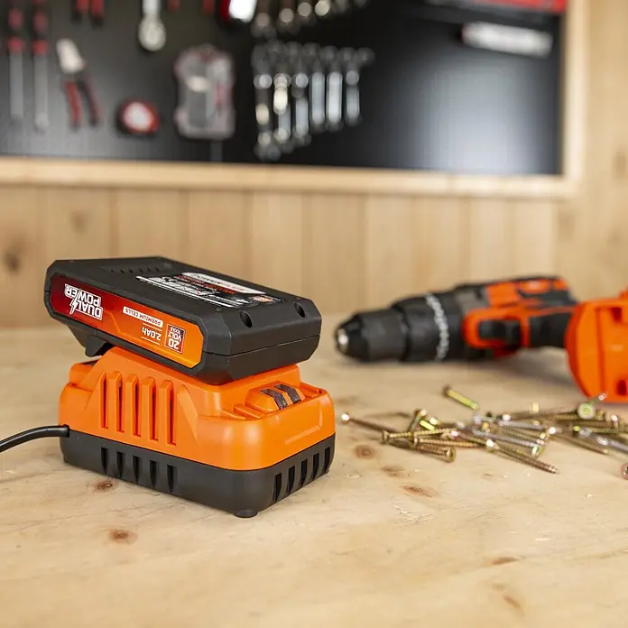 PowerPlus Tijeras para podar de batería + Motosierra - 20 V, 1 batería, 2 Ah, Grosor de rama máx.: 30 mm - Batería de taladro inalámbrico negra y naranja cargando en su cargador a juego, con taladro y tornillos borrosos al fondo.