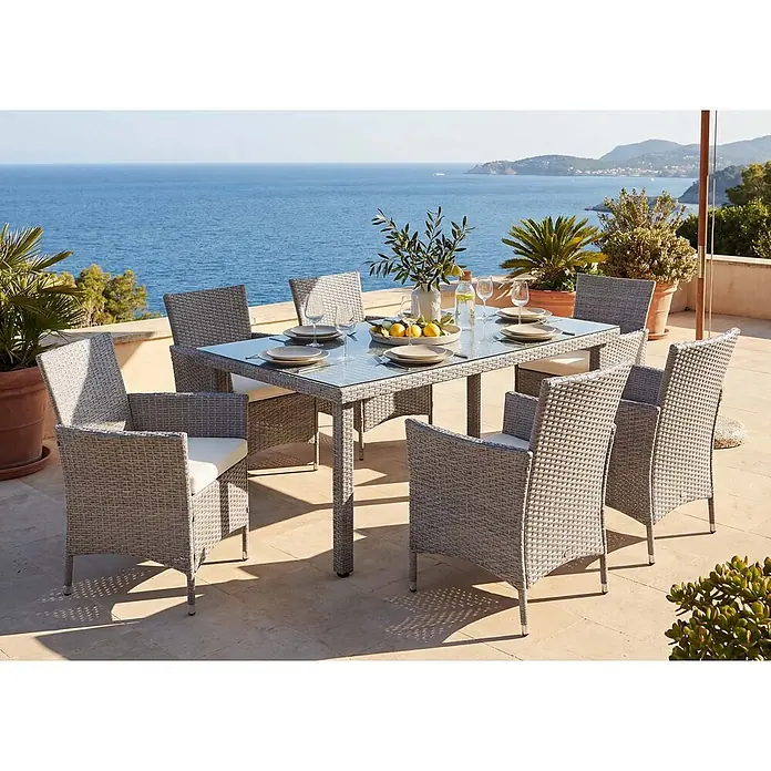 Mendler Gartenmöbel-Set - 7 -tlg., Polyrattan, Grau, Kissen creme, Grau, Kissen creme - Graues Polyrattan-Gartenset mit Glastisch und Stühlen mit weißen Kissen, auf einer Terrasse mit Meerblick.